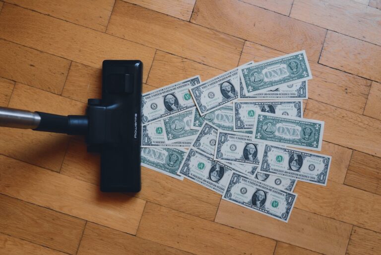 A vacuum cleaner on a wooden floor with scattered one-dollar bills in front of it, symbolizing the art of budgeting and efficient financial planning.