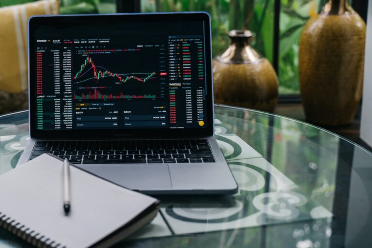 A laptop showcasing cryptocurrency trading charts and data rests on a glass table next to a spiral notebook and pen, tools essential for any financial planner. Elegant vases and lush greenery enhance the sophisticated backdrop.