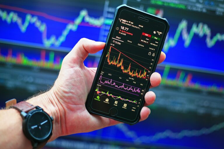 A hand holds a smartphone displaying stock market graphs and data, set against a backdrop of larger financial charts on screens, highlighting the tools a savvy financial planner might use to manage credit and budget effectively.