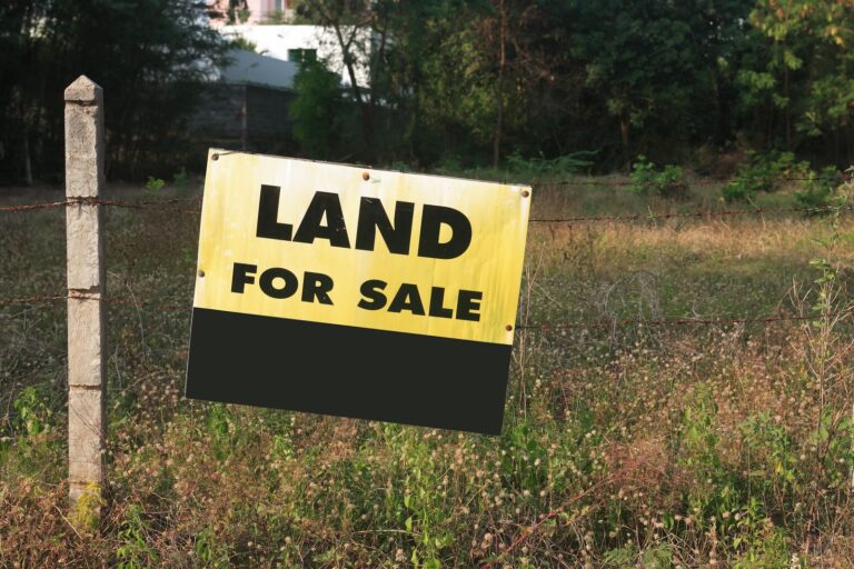 A "Land for Sale" sign is attached to a wire fence, hinting at an ideal investment opportunity for any budget, with lush grass and trees in the background.