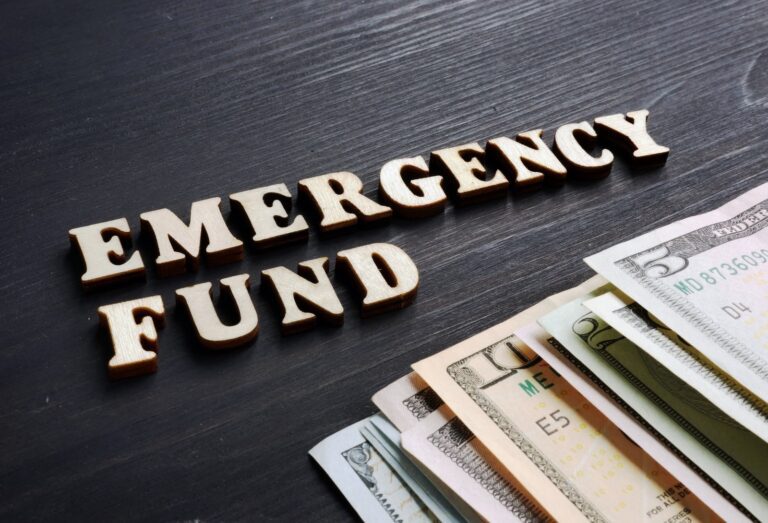 Wooden letters spell "EMERGENCY FUND" on a dark surface beside assorted US dollar bills, highlighting the importance of careful budgeting.