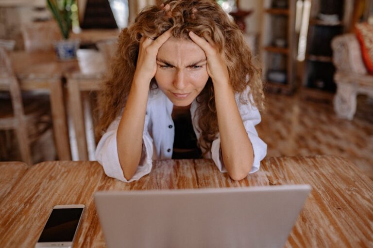 A financial planner with long curly hair looks frustrated while sitting at a table, trying to manage a budget on their laptop. A smartphone is placed on the table.