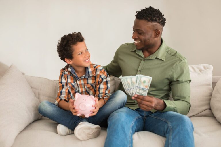 A man and child sit on a couch, smiling at each other. The man, a financial planner, holds dollar bills, while the child holds a pink piggy bank, learning about budgeting in this joyful moment.