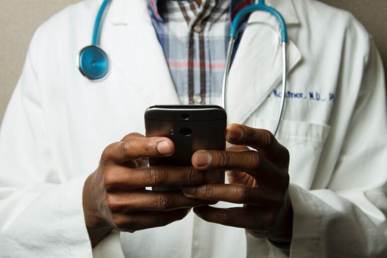 A person in a white lab coat uses a smartphone, perhaps checking a budget app. A stethoscope is draped around their neck, highlighting their dual focus on health and finances.