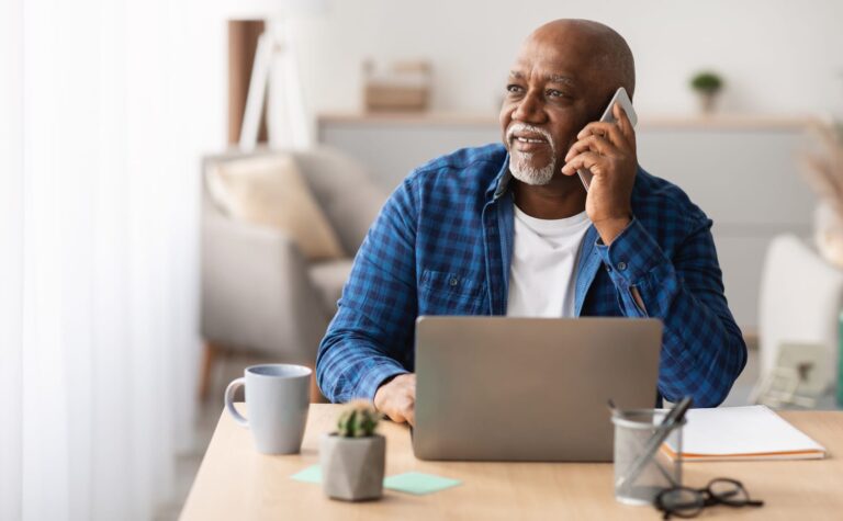 Senior Black Man At Laptop Talking On Phone Looking Aside Communicating For Work Sitting At Table In Office. Entrepreneur Chatting By Cellphone At Workplace. Mobile Communication