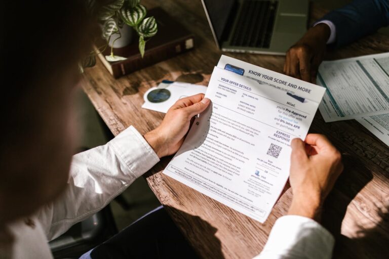 The image shows a person's hands reviewing a document titled "Know Your Score and More" that appears to contain information about business tradelines. The document has details about the person's financial profile, potential offers, and other related data. The setting includes a potted plant and other papers on a wooden table, suggesting a professional or business context.