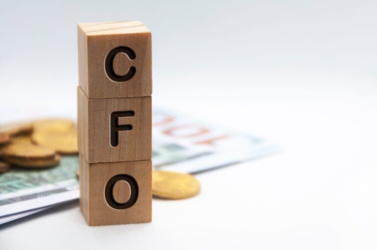 Wooden blocks spelling "CFO" sit prominently among coins, while a blurred banknote looms in the background, capturing the attention of CFO recruiters searching for financial leaders.