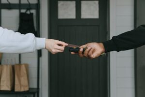 Two hands exchanging a credit card in front of a door, with shopping bags subtly hinting at hidden fees lurking in the background.