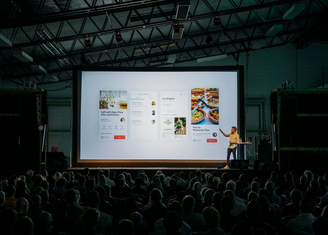 A speaker presents a slideshow to a large seated audience in a dimly lit auditorium. The slide showcases mobile app interface designs enhanced by AI tools, featuring captivating images and text.