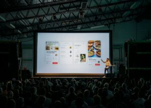 A speaker presents a slideshow to a large seated audience in a dimly lit auditorium. The slide showcases mobile app interface designs enhanced by AI tools, featuring captivating images and text.