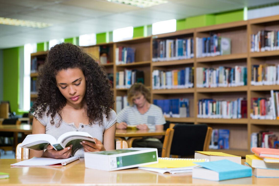 A person enjoys reading a book at a library table, surrounded by shelves of books, with another individual seated further away. The cozy atmosphere is ideal for students, who might even take advantage of exclusive library discounts.