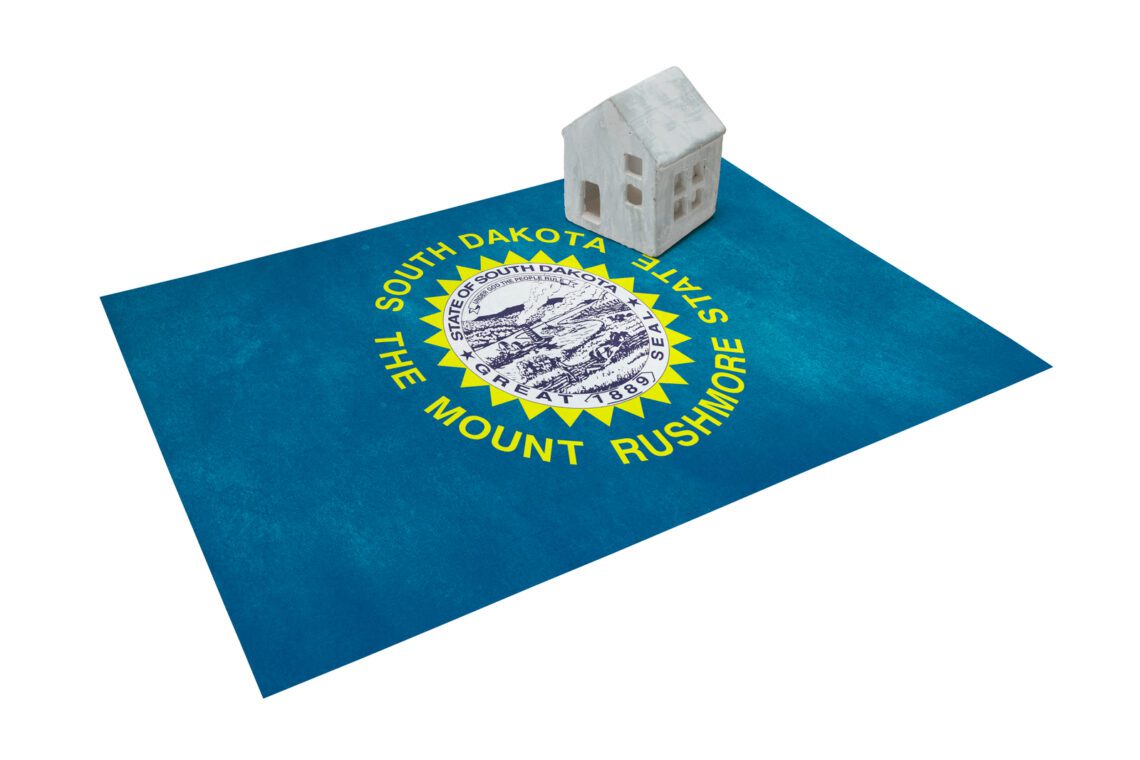 A small white house model rests on a South Dakota state flag, showcasing the Mount Rushmore emblem—a perfect symbol for those considering buying a home in South Dakota.