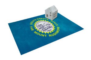 A small white house model rests on a South Dakota state flag, showcasing the Mount Rushmore emblem—a perfect symbol for those considering buying a home in South Dakota.