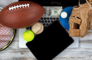 A baseball glove, football, tennis ball, racket, ping pong ball, dollar bill, and laptop are arranged on a wooden surface, painting a picture of sports betting enthusiasts preparing for the action both on the field and online.