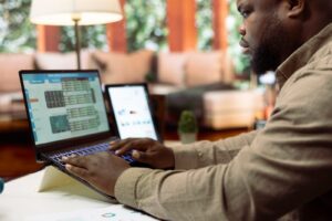 A man sits at a desk in his living room, scrutinizing data on his laptop as he plans the small business budget for 2025. Nearby, another device and documents are within reach, aiding his meticulous preparation.