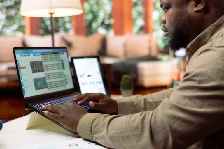 Categories 20 A man sits at a desk in his living room, scrutinizing data on his laptop as he plans the small business budget for 2025. Nearby, another device and documents are within reach, aiding his meticulous preparation.