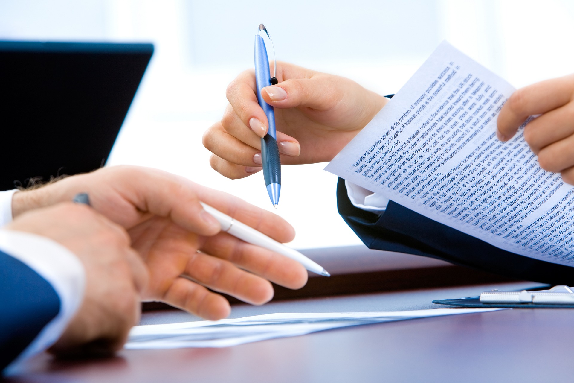 Two individuals in business attire are discussing a document, perhaps the role of a financial advisor in retirement planning. One points at the paper with a pen while the other holds a similar one, with a laptop partially visible in the background.