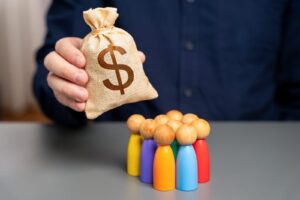 A person holds a burlap sack with a dollar sign, symbolizing small business funding, above colorful wooden figurines on a table.