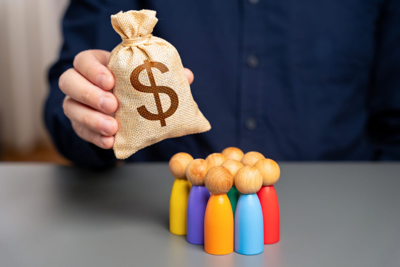 A person holds a burlap sack with a dollar sign, symbolizing small business funding, above colorful wooden figurines on a table.