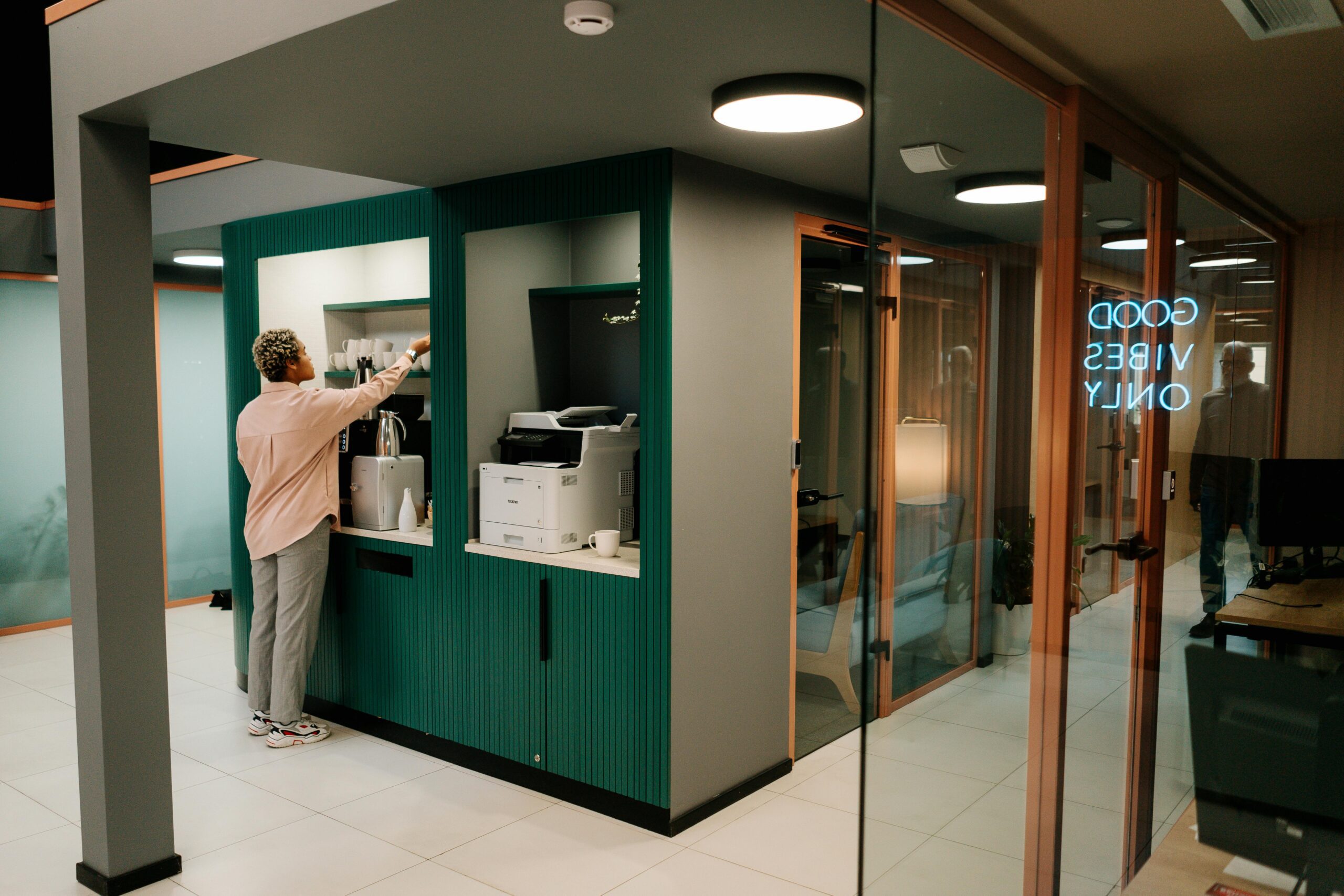 Amidst the office space's cozy glow of a neon "GOOD VIBES ONLY" sign, a person reaches for items on the shelf, while nearby, a printer hums with activity—hints of business expansion in progress.