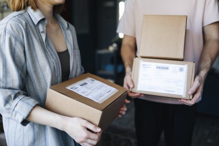 Two people hold cardboard boxes with shipping labels in a room, poised for delivery as part of their small business shipping operations.