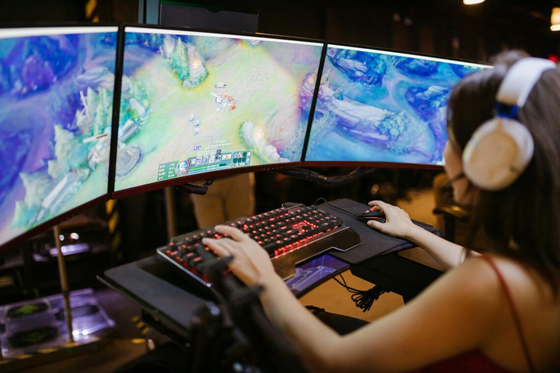 A person immerses themselves in low-cost online entertainment options, wearing headphones and playing a video game on a triple-monitor setup with a mechanical keyboard and mouse.