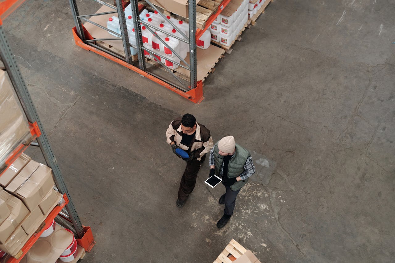 Two people stand in a warehouse aisle, one holding a tablet running an inventory management app. Shelves with various packages are visible.