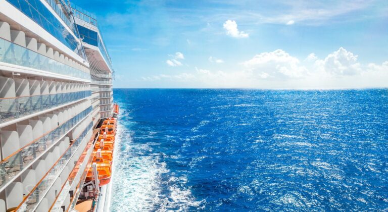 A cruise ship sails through the bright blue sea under a clear sky, with lifeboats visible along its side.