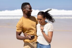 A couple stands on a beach, smiling and laughing as they share a moment. The man holds a smartphone, perhaps checking how to choose a financial advisor, while the waves gently lap in the background.