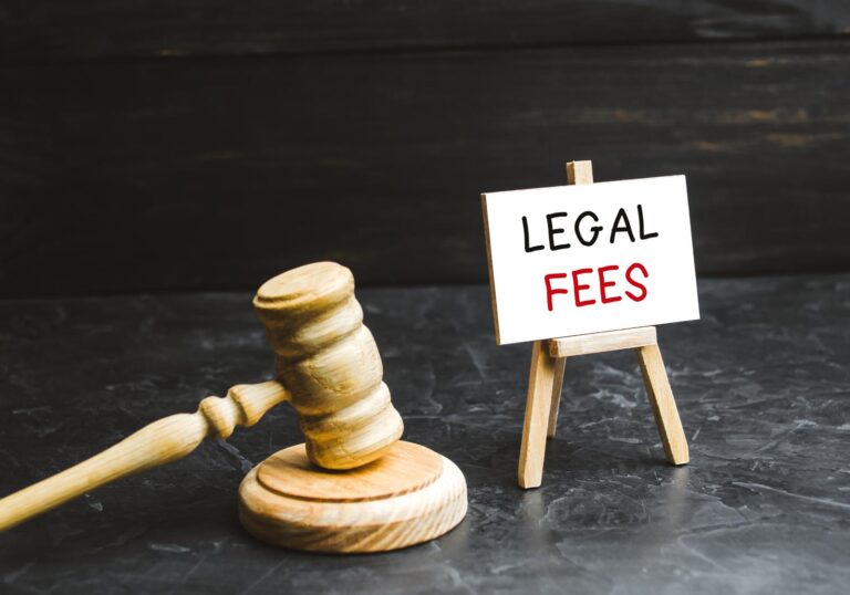 A wooden gavel rests beside a small easel with a sign that reads "Lawyer Fees" on a dark surface.