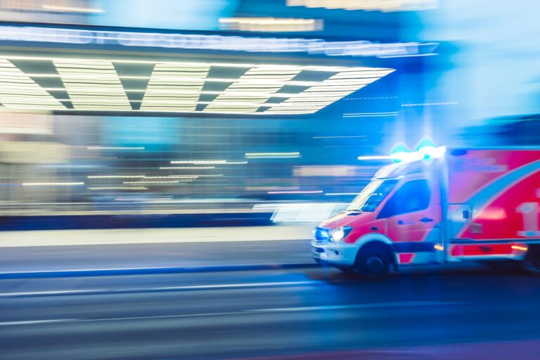 A blurred image of a red and white ambulance with flashing lights speeds down a city street, reminding us of the urgent care we might rely on, highlighting concerns about healthcare costs in retirement.