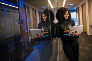 An employee with curly hair uses a laptop in the hallway, demonstrating security awareness as they stand beside a server room with glass walls.