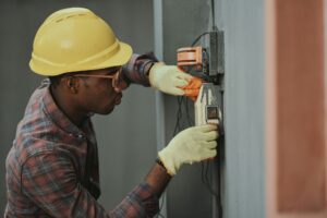 A person wearing a yellow hard hat and gloves diligently works with electrical wires on a gray wall, ensuring smooth business electricity operations.