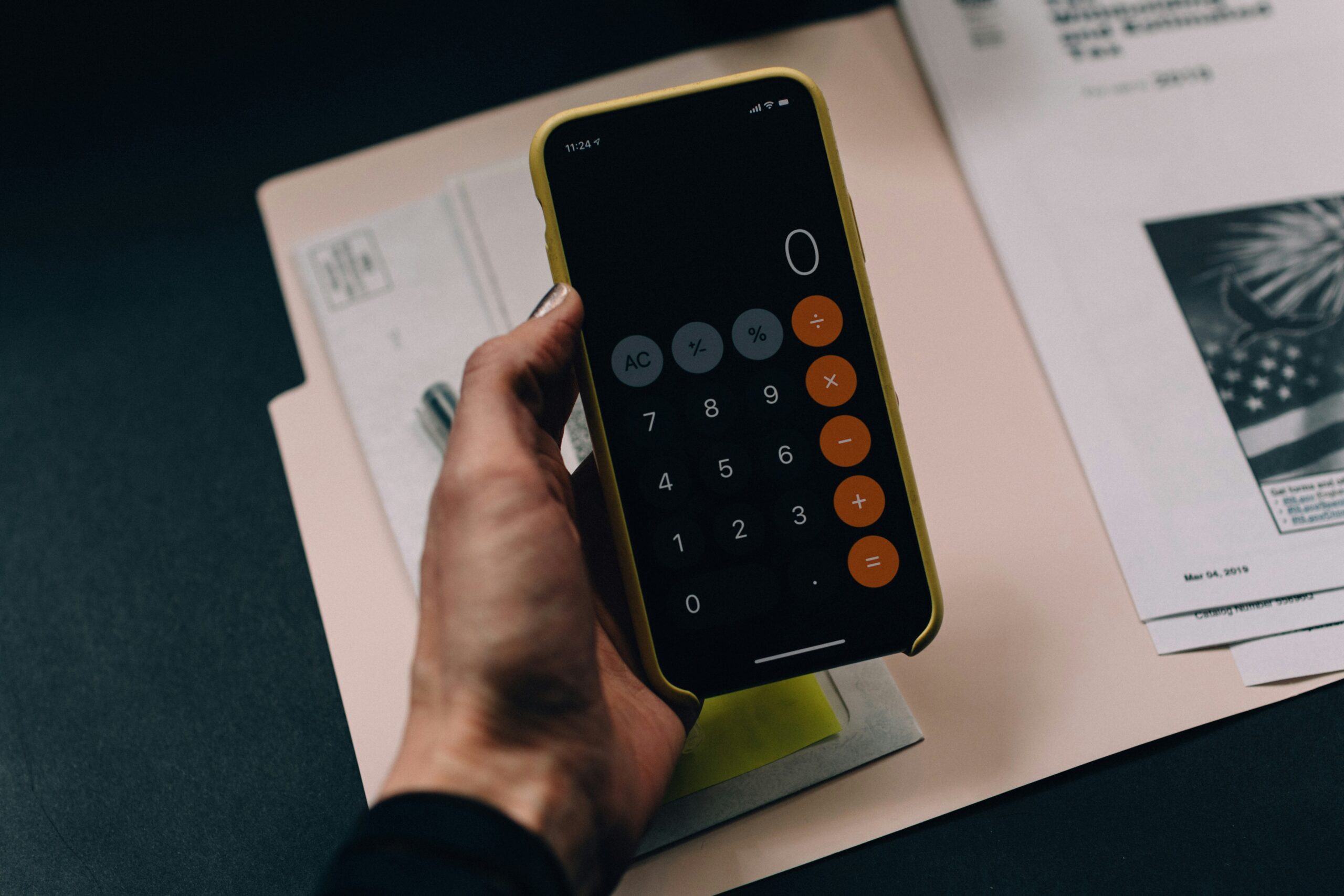 A hand holds a smartphone displaying a calculator app, aiding in financial decluttering, with scattered papers and documents in the background.