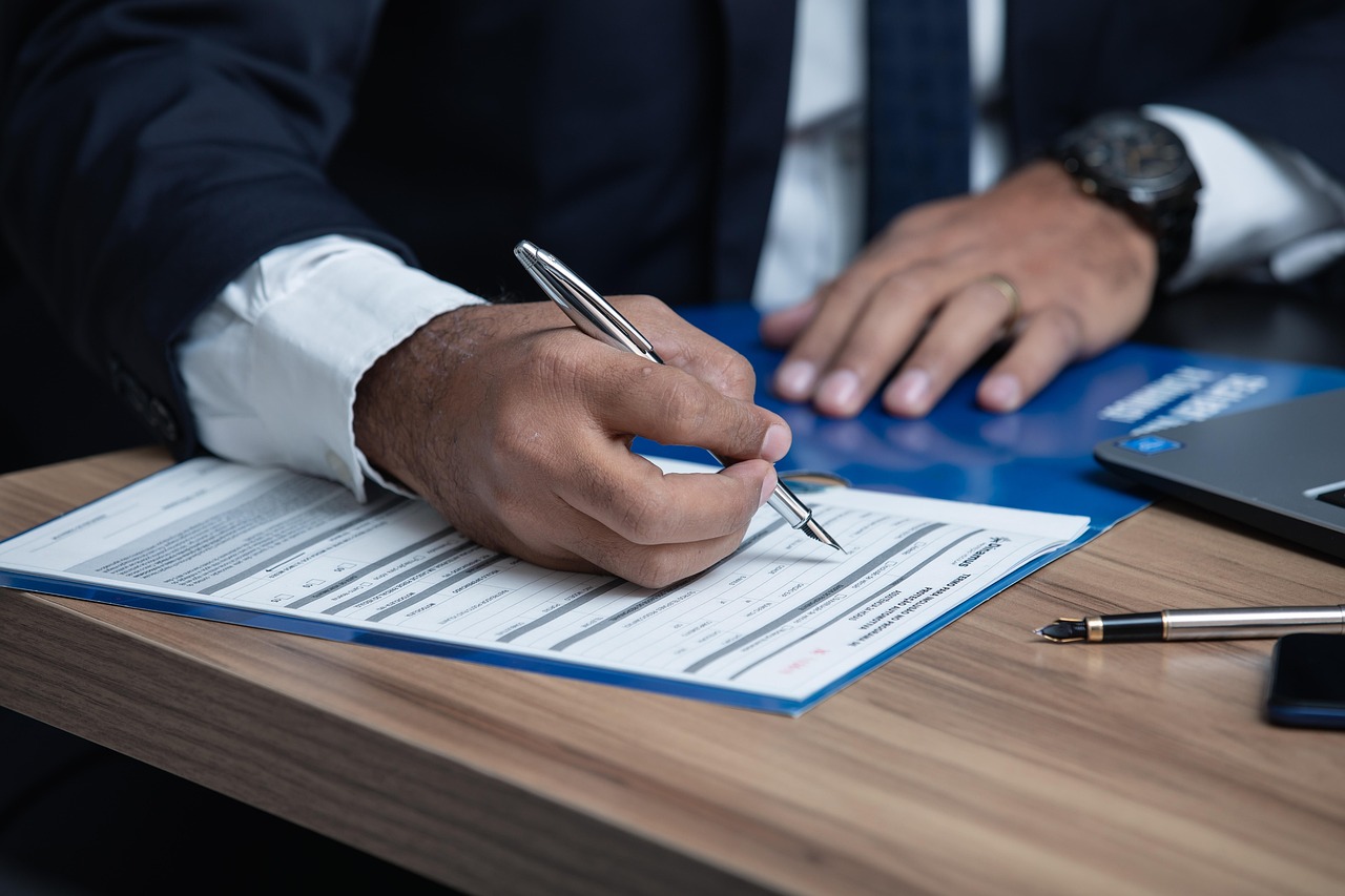 A person in a suit is meticulously signing a document with a silver pen. On the wooden table, alongside a laptop and blue folder, lies evidence of precise detail often demanded by legal staffing services.