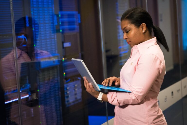 A person in a pink shirt examines used servers, utilizing a laptop amidst the blue-lit server racks of the room.