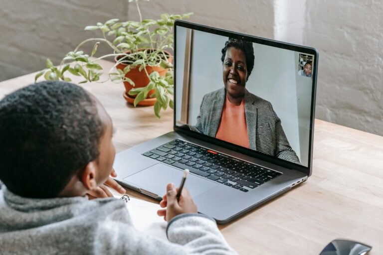 A person engages in a video call on a laptop, connecting with an online tutor. On the table beside them are a potted plant and a notebook.