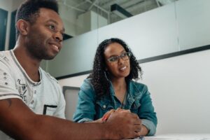 Two people sit at a table in an office setting, gazing intently off-camera. The person on the left wears a crisp white shirt, while the individual on the right sports a casual denim jacket. They could be seeking advice on how to choose a financial advisor.