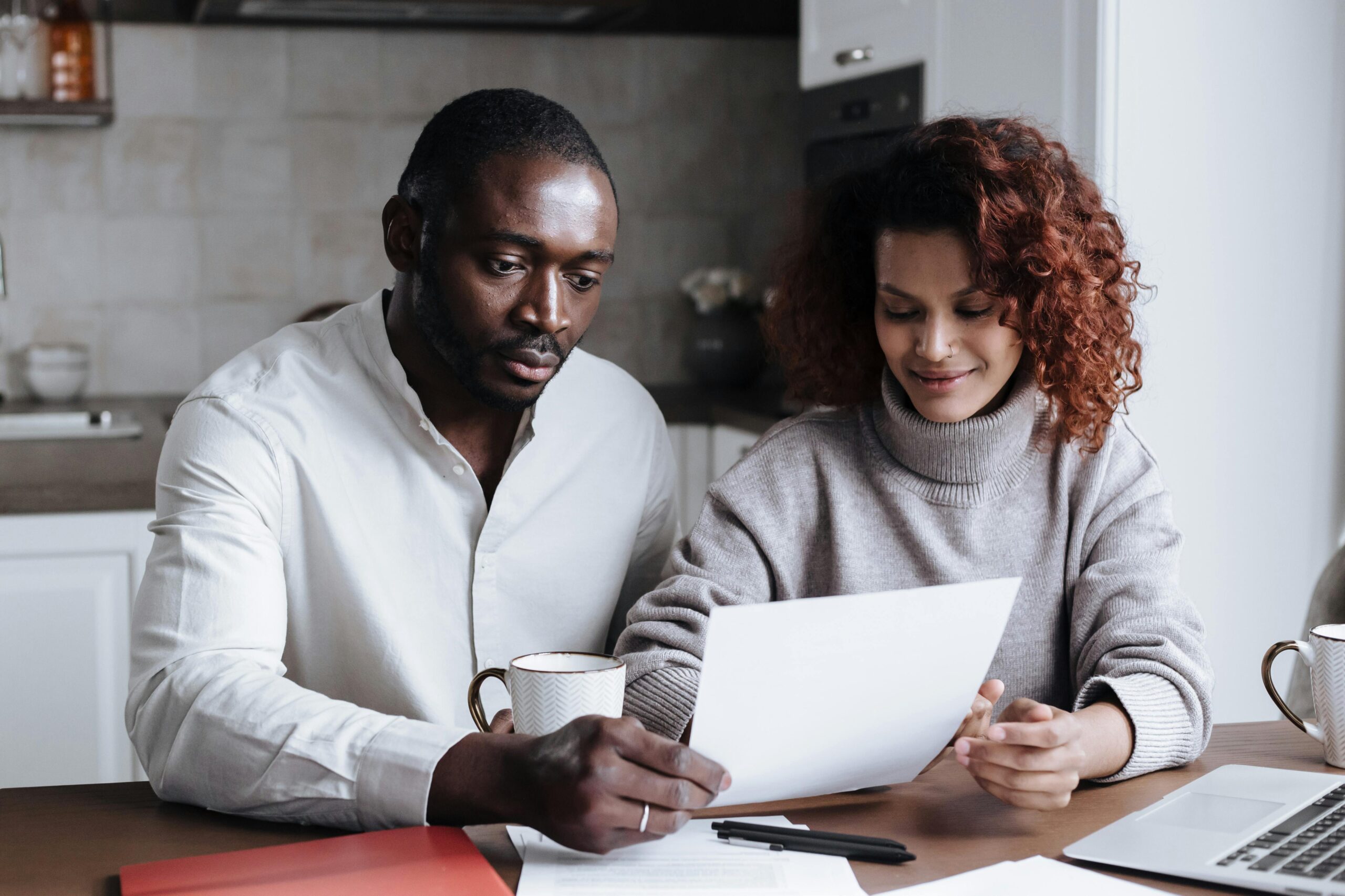 A man and woman sit at a table, reviewing a document together. With coffee mugs steaming beside them and a laptop open, they're discussing the best approach to find a financial advisor.