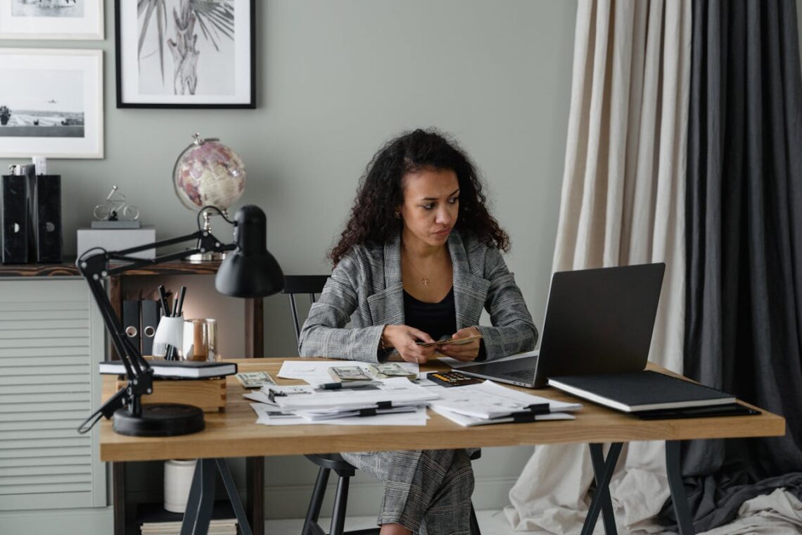 The person, in the midst of an executive relocation, is seated at a desk with a laptop and paperwork, holding a piece of paper. The desk features a lamp, globe, and stationery. Wall art and curtains set the backdrop for this transitional workspace.