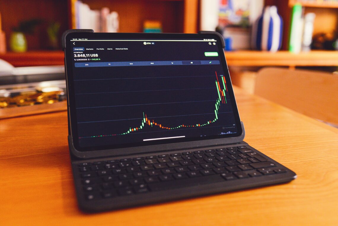 A tablet showcasing a vibrant crypto trading candlestick chart rests on a wooden table, with shelves lining the background.