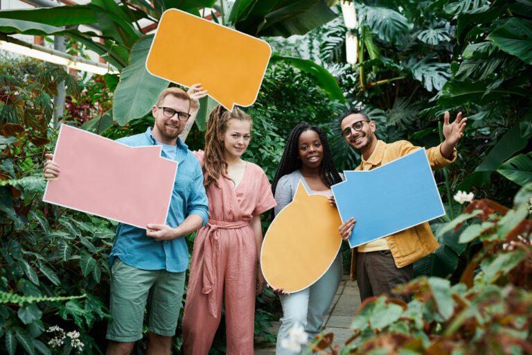 Four people stand in a greenhouse, each holding a large colorful speech bubble cutout—an inspiring way to add speech bubbles to travel photos, surrounded by vibrant green plants.