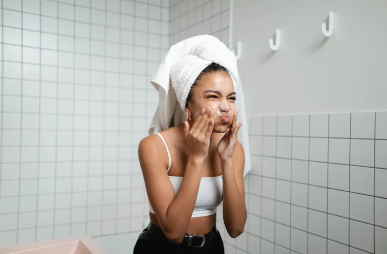 A woman with a towel wrapped around her head washes her face in a tiled bathroom, standing near a pink sink and empty wall hooks, reflecting on the lifetime cost of women’s healthcare.