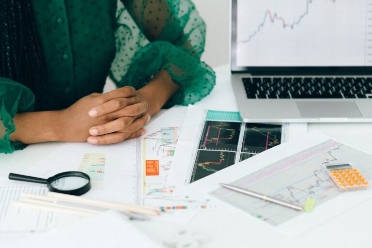 A person sits at a desk with folded hands, surrounded by financial charts, graphs, a calculator, a magnifying glass, and a laptop displaying a line graph—reflecting thoughtful September money moves.