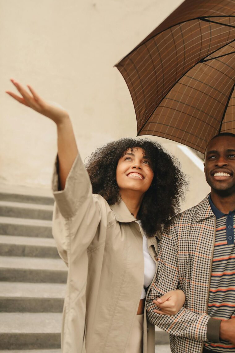A woman smiles and reaches out her hand while standing next to a man under a brown umbrella near stairs, capturing a moment of connection that feels as effortless as using Self Cash.