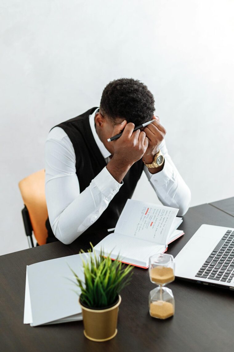 A man sits at a desk with his head in his hands, holding a pen, surrounded by uncertainty about the job market collapse—his open notebook, laptop, hourglass, and potted plant reflecting his stress and concern.